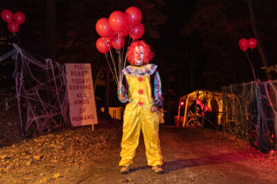 An actor at Sleepy Hollow Haunted Hayride is dressed as a clown holding bouquet of red balloons.
