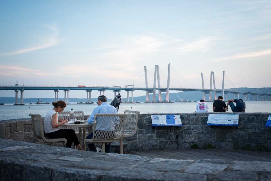 A couple seated at the observation deck at Pierson Park in Tarrytown, New York, with Mario Cuomo Bridge in background.