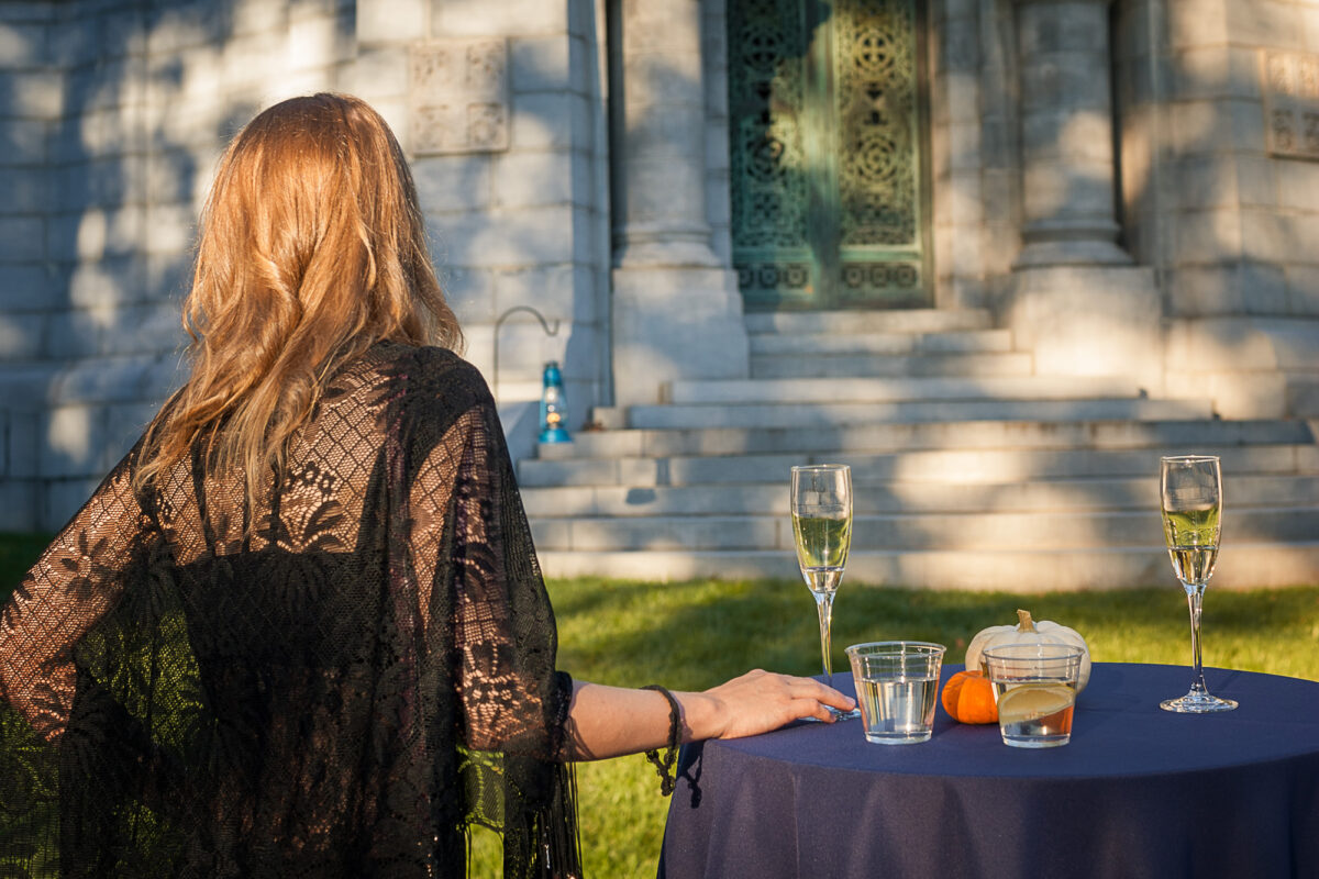 A woman in a black lace shawl stands with her back to the camera at a cocktail table, gazing toward the ornate bronze door and marble steps of a mausoleum lit by late-afternoon sun. The table holds two champagne flutes, two cocktails, and small pumpkins, at Champagne Cocktails with the Dead at Sleepy Hollow Cemetery.