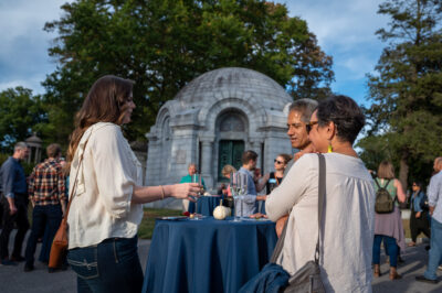 Guests mingle with champagne flutes at cocktail tables outside a domed marble mausoleum at golden hour. In the foreground, a woman in a cream blouse chats with a couple beside a blue-clothed table set with champagne and a small white pumpkin, at Champagne Cocktails with the Dead at Sleepy Hollow Cemetery.