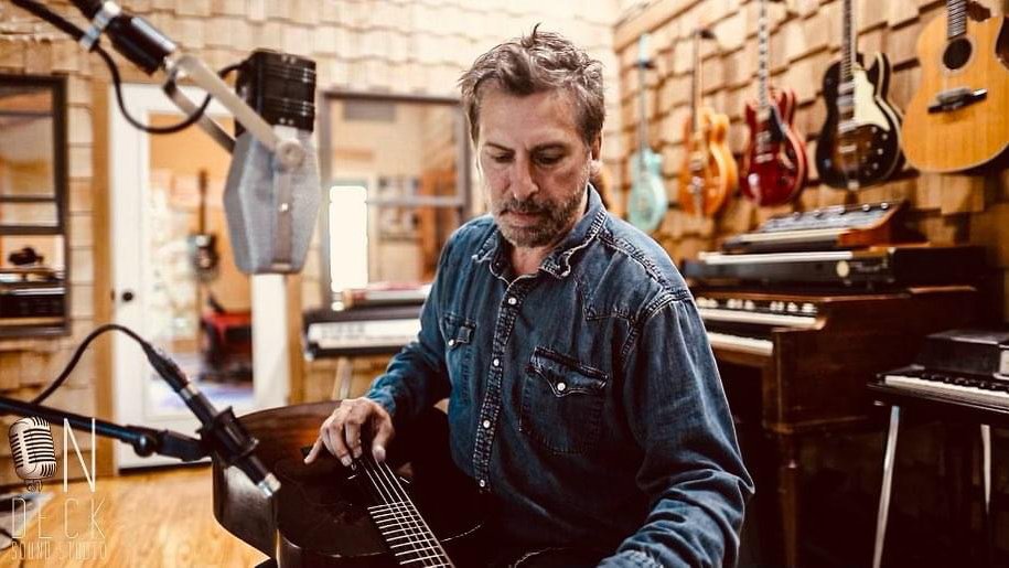 Musician Joe Crookston poses in a recording studio with a guitar in hand.